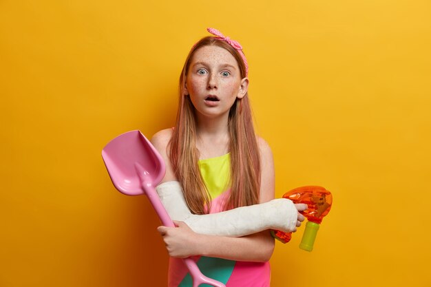 Concepto de niños, ocio y juegos. Una niña sorprendida juega con una pistola de agua, sostiene una pala de arena, se divierte en la playa durante el verano, impresionado al ver algo increíble usa un traje de baño en el brazo lesionado