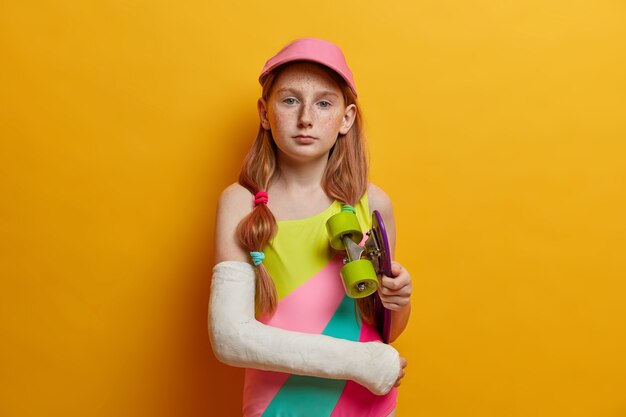 Concepto de niños, pasatiempos y recreación. Grave pelirroja pecosa posa con patineta, se traumatizó después de montar a alta velocidad, le gustan los deportes extremos. El patinador infantil sostiene el longboard debajo del brazo