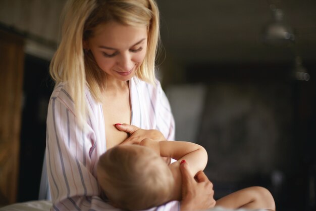 Concepto de niños recién nacidos, infancia, cuidado de niños y maternidad. Madre joven soñolienta con cabello rubio desordenado amamantando a su hijo por la noche. Bastante mamá dando mama a un niño de siete meses