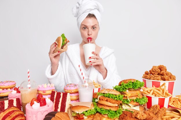 Concepto de nuttrition malsano. Ama de casa con manicura roja y labios en toalla de albornoz doméstico en la cabeza bebe refrescos come comida chatarra