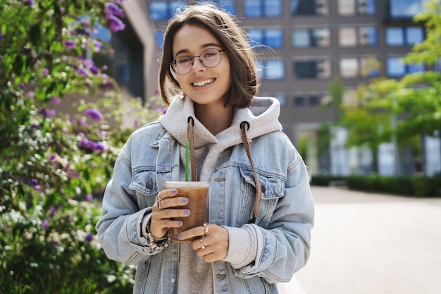 Concepto de ocio y actividades al aire libre en el estilo de vida Retrato de una chica queer feliz con chaqueta vaquera y gafas paseando por la ciudad disfrutando de la primavera bebiendo café con leche helado y una cámara sonriente