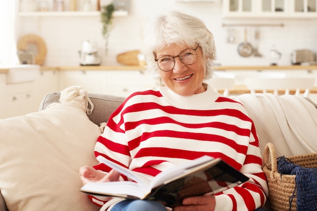 Concepto de ocio, autoeducación, afición y jubilación. Imagen de buena mujer senior madura en suéter a rayas y elegantes gafas disfrutando de la lectura en la sala de estar, sonriendo con alegría