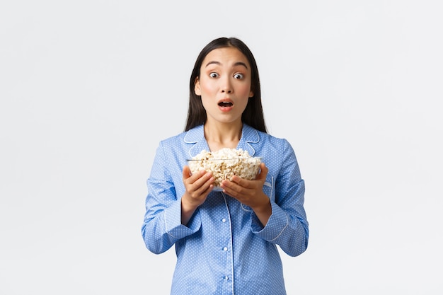 Concepto de ocio en casa, pijamada y fiesta de pijamas. Asombrada linda chica asiática en pijama, sosteniendo un tazón de palomitas de maíz y mandíbula caída, jadeando mientras mira la televisión viendo una escena de película interesante, fondo blanco.