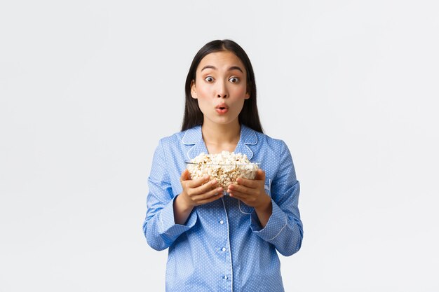 Concepto de ocio en casa, pijamada y fiesta de pijamas. Chica asiática emocionada e intrigada en pijama mirando con diversión e interés en la pantalla del televisor, viendo películas y comiendo palomitas de maíz, fondo blanco.