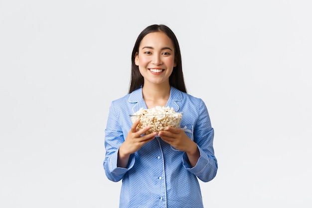 Concepto de ocio en casa, pijamada y fiesta de pijamas. Sonriente niña asiática complacida disfrutando los fines de semana en la cama con palomitas de maíz, comiendo y viendo películas en pijama, de pie fondo blanco.