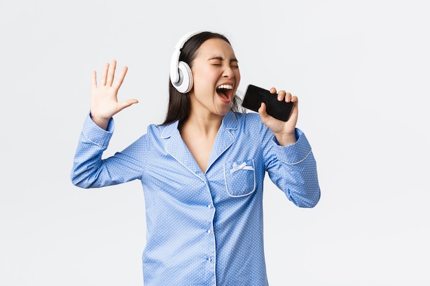 Concepto de ocio, fines de semana y estilo de vida en el hogar. Chica asiática emocionada y despreocupada en pijama, jugando la aplicación de karaoke en el teléfono inteligente, cantando canciones en el teléfono móvil con auriculares, fondo blanco.