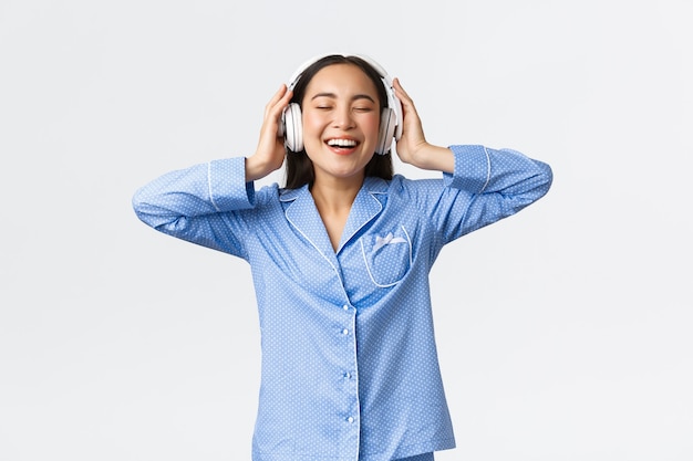Concepto de ocio, fines de semana y estilo de vida en el hogar. Feliz mujer asiática complacida en pijama disfrutando de una calidad de sonido impresionante en auriculares nuevos, bailando en pijama y escuchando música.