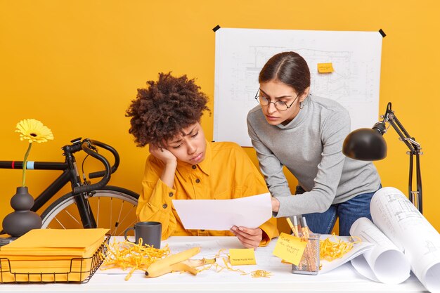 Concepto de ocupación de trabajo de personas. Tristes mujeres profesionales diversas intentan decidir el problema con errores en los bocetos tienen algunas dificultades planteadas en el espacio de coworking discutir el plan de negocios centrado en los documentos