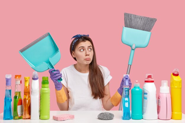 Concepto de orden y limpieza. Atractiva mujer de cabello oscuro usa diadema, tiene aspecto molesto, lleva escoba y pala, limpia la casa sola