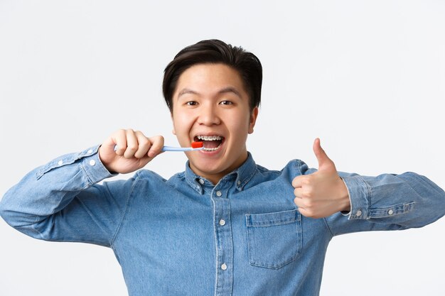 Concepto de ortodoncia, cuidado dental e higiene. Primer plano de hombre asiático feliz satisfecho cepillarse los dientes con tirantes, sosteniendo el cepillo de dientes y mostrando el pulgar hacia arriba en señal de aprobación, fondo blanco.