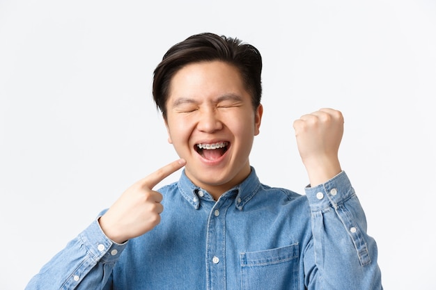 Concepto de ortodoncia y estomatología. Primer plano de un chico asiático emocionado y feliz regocijándose con los nuevos apoyos de los dientes, apuntando a la boca y sonriendo, bomba de puño, triunfando sobre el fondo blanco