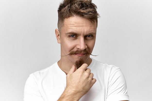 Concepto de peluquería, moda y estilo. Imagen de hombre de aspecto moderno con elegante corte de pelo, bigote y barba de chivo mirando a la cámara con una sonrisa misteriosa, mirando por debajo de sus espesas cejas