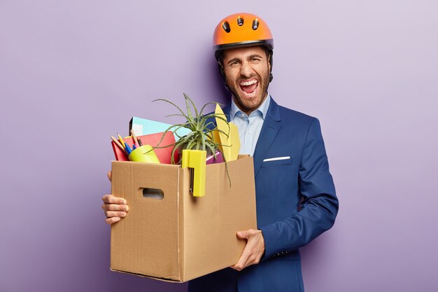 Concepto de pérdida de empleo. Ingeniero hombre molesto despedido del trabajo, lleva una caja de cartón con material de oficina personal, sonríe