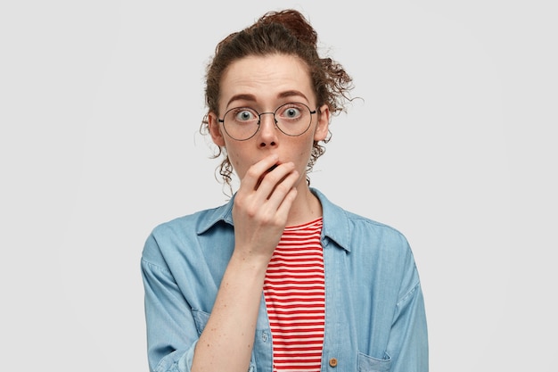 Concepto de personas y asombro. Mujer joven aturdida y pecosa con mirada asustada, mantiene la mano en la boca, cotillea con un amigo, jadea de miedo y asombro, usa anteojos redondos, camisa informal