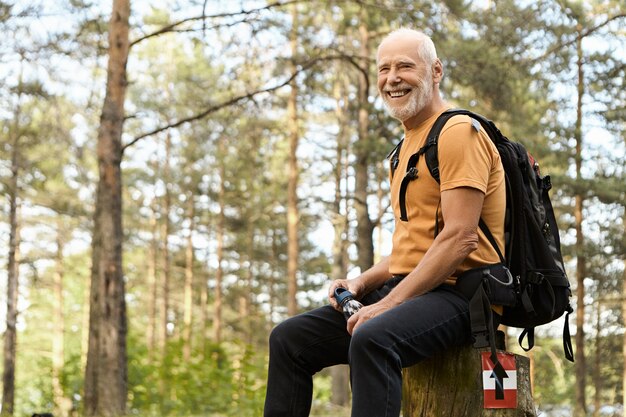 Concepto de personas, aventuras, viajes y estilo de vida activo y saludable. Alegre anciano enérgico senderismo con mochila en el bosque, descansar sobre tocón, agua potable con pinos en