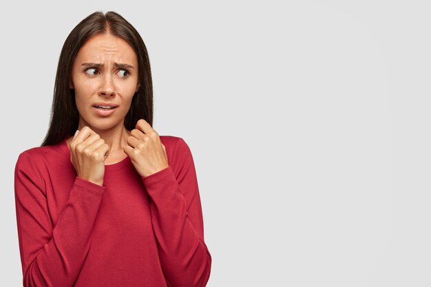 Concepto de personas y aversión. Aspecto femenino atractivo disgustado con expresión facial negativa asustada, vestida con suéter rojo, se encuentra contra la pared blanca