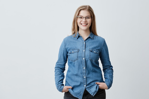 Concepto de personas, belleza y estilo de vida. Atractiva mujer rubia sensual con gafas y amplia sonrisa vestida con camisa vaquera sonriendo ampliamente estando feliz de conocer a su mejor amiga. Alegre agradable hembra