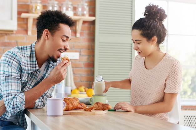 Concepto de personas, cocina y degustación. Pareja familiar almuerza en una acogedora cocina: hombre barbudo de piel oscura come delicioso croissant dulce