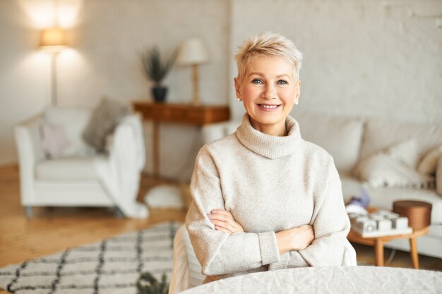 Concepto de personas, comodidad, domesticidad y temporada. Encantadora hermosa mujer jubilada pasar tiempo libre en el interior de su casa con una sonrisa de confianza, manteniendo los brazos cruzados sobre el pecho