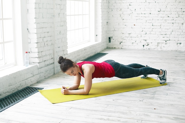 Concepto de personas, deportes, fitness, fuerza y resistencia. Chica atlética morena con estilo en ropa deportiva haciendo ejercicio en el espacioso salón junto a la ventana, haciendo tabla de codo en la estera de yoga verde sobre piso de madera