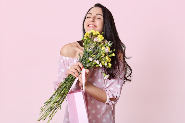 Concepto de personas, disfrute y felicidad. Mujer de cabello oscuro positiva con cabello negro, abraza flores, lleva bolsa de regalo, posa en rosa claro. La hembra se alegra el 8 de marzo.