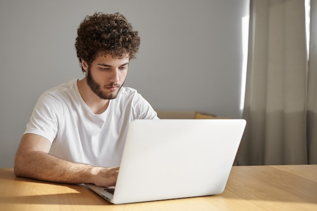 Concepto de personas, dispositivos electrónicos y tecnología. Foto sincera de un profesional independiente masculino joven guapo serio que usa wifi gratuito en la computadora portátil mientras trabaja de forma remota desde la oficina en casa, con una mirada enfocada