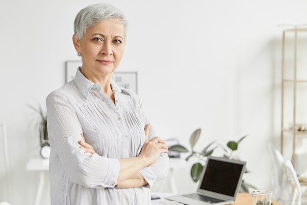 Concepto de personas, edad, tecnología y trabajo. Atractiva ejecutiva de mediana edad seria con peinado corto de duendecillo de pie en la oficina con los brazos cruzados sobre el pecho, su postura expresando confianza