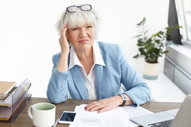 Concepto de personas, edad, trabajo, estrés y salud. Imagen de empresaria de pelo gris disgustada frunciendo el ceño, tocando la cabeza para aliviar el dolor debido al dolor de cabeza, trabajando demasiado, estudiando papeles en la oficina