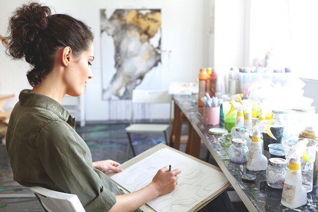 Concepto de personas, educación y creatividad. Perfil de mujer joven con cabello rizado en cola de caballo aprendiendo a dibujar y dibujar mientras participa en un taller de arte o clase