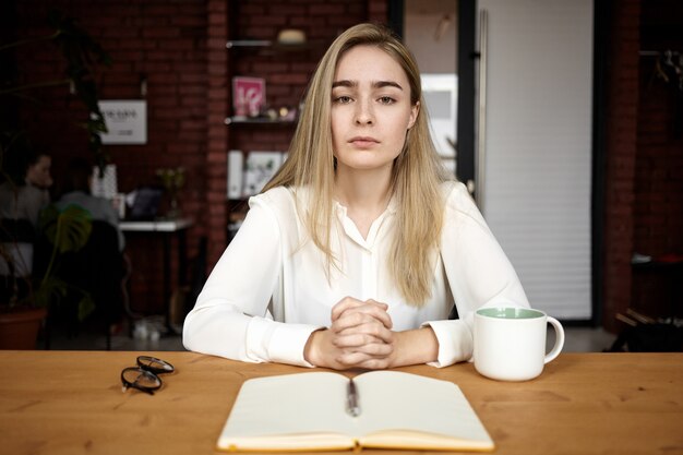 Concepto de personas, educación, trabajo y autónomo. Elegante joven autónoma o estudiante chica sentada a la mesa en el café, tomando café, esperando a un amigo o cliente, abra el cuaderno frente a ella