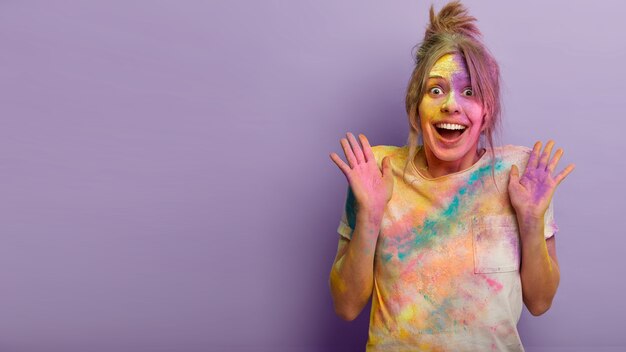 Concepto de personas, emociones y vacaciones. Una mujer europea feliz y emotiva levanta las palmas de la felicidad, no puede evitar los sentimientos positivos, impresionado por la celebración del Festival de los colores de Holi en el extranjero