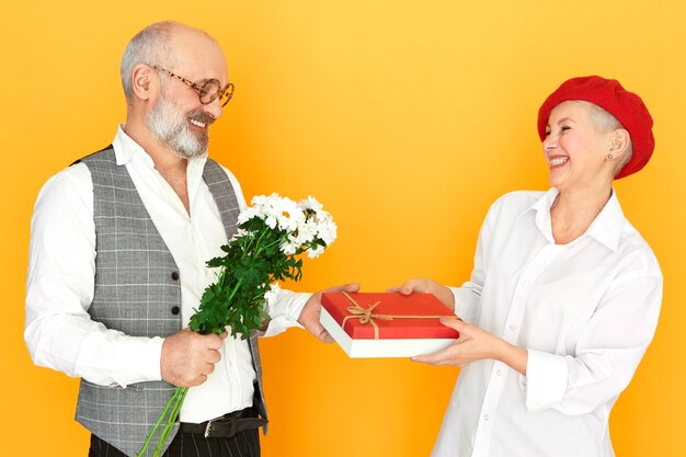 Concepto de personas, envejecimiento, citas y romance. Vista lateral del apuesto hombre senior elegante en vasos con ramo de flores de campo y caja de chocolate, dando presente a su atractiva fecha de mujer madura