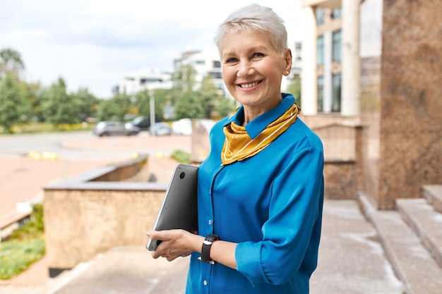 Concepto de personas, envejecimiento, estilo de vida urbano, carrera y tecnología. Elegante elegante empresaria de mediana edad con portátil posando fuera del edificio de oficinas, yendo a una reunión de negocios, sonriendo a la cámara