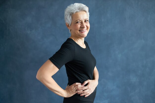 Concepto de personas, envejecimiento, madurez y estilo de vida. Imagen de estudio de mujer madura alegre y llena de alegría en top apretado negro haciendo actividad física, posando aislado con las manos en la cintura, riendo