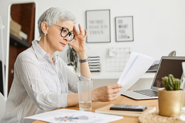 Concepto de personas, envejecimiento, tecnología y profesión. Grave mujer caucásica de 50 años con elegantes lentes y camisa de seda leyendo un contrato mientras trabaja en el escritorio, sentado frente a una computadora portátil abierta