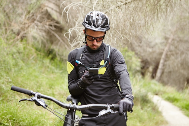 Concepto de personas, estilo de vida activo, deportes y tecnología. Jinete masculino serio con equipo de protección usando internet en el teléfono móvil durante un pequeño descanso mientras monta en bicicleta con motor al aire libre