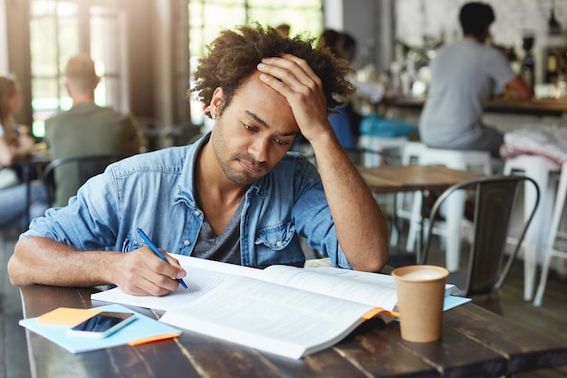 Concepto de personas, estilo de vida, aprendizaje, conocimiento y educación. Retrato de atractivo estudiante afroamericano centrado ocupado trabajando en papel del curso, tomando notas del libro de texto en el cuaderno