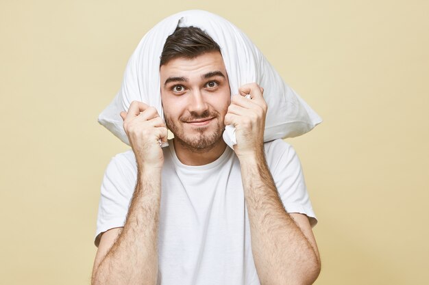 Concepto de personas, estilo de vida, descanso, salud y recreación. Apuesto joven sin afeitar en pijama blanco cubriendo las orejas con la almohada, sonriendo, porque no escucha el ronquido de su esposa dormida