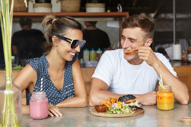 Concepto de personas y estilo de vida. Dos amigos que tienen una conversación agradable disfrutando de comida sabrosa durante el almuerzo. Joven comiendo papas fritas y hablando con su atractiva novia en elegantes gafas de sol