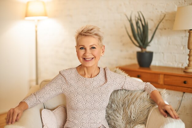 Concepto de personas, estilo de vida y edad madura. Imagen horizontal de buena mujer confiada con el pelo corto teñido relajándose en casa, sentado cómodamente en un sillón, sonriendo ampliamente