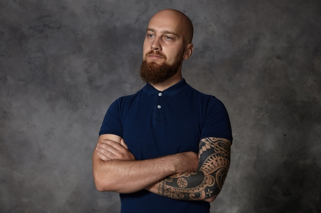 Concepto de personas y estilo de vida. Hombre tatuado de moda guapo aislado con barba difusa y cabeza rapada posando en el interior con los brazos cruzados, con expresión facial segura