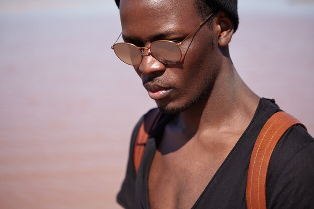 Concepto de personas, estilo de vida y moda. Retrato de joven guapo modelo masculino afroamericano de moda con mochila de cuero con elegante camiseta negra y gafas de sol posando al aire libre