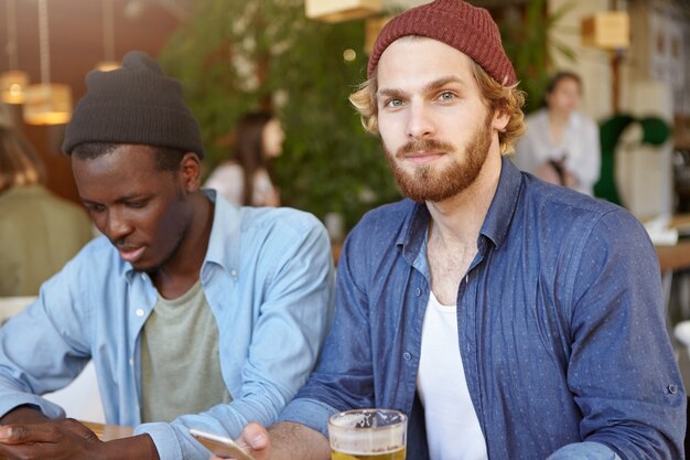 Concepto de personas, estilo de vida moderno, amistad, relaciones y tecnologías. Dos hombres guapos y elegantes que se relajan en el café o en el bar, beben cerveza y se divierten, usando wifi gratis en teléfonos móviles