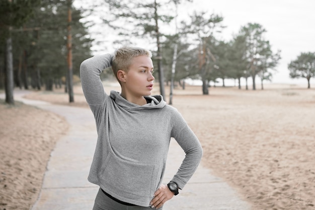 Concepto de personas, estilo de vida, ocio y actividad. Retrato de deportista rubia elegante con capucha caminando afuera en el parque público, con mirada pensativa. Chica guapa con entrenamiento atlético al aire libre