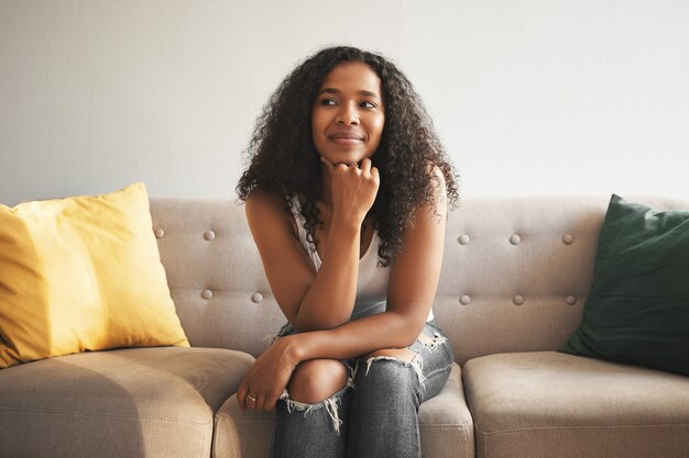 Concepto de personas y estilo de vida. Retrato de alegre hermosa joven afroamericana vistiendo elegantes jeans pasar tiempo en casa, sentado en un cómodo sofá, mirando a otro lado con una sonrisa de ensueño