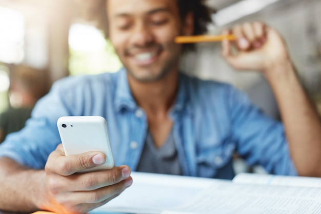 Concepto de personas, estilo de vida, tecnología y comunicación. Apuesto estudiante de piel oscura con barba y camisa azul usando un teléfono celular, navegando por las noticias a través de las redes sociales, riéndose de los memes