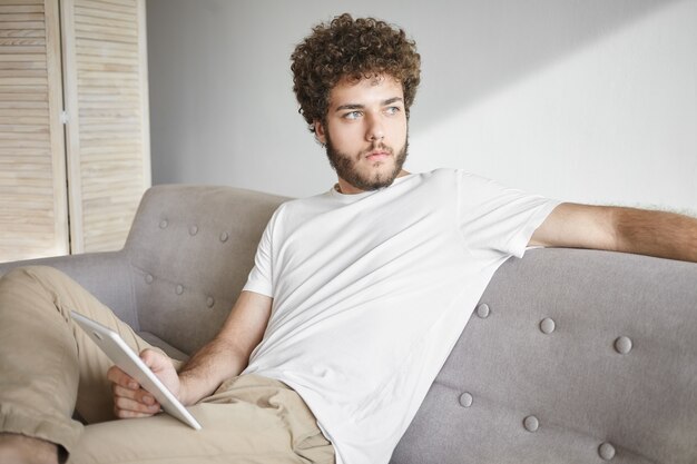 Concepto de personas, estilo de vida, tecnología y comunicación. Blogger masculino joven atractivo con barba difusa y cabello rizado con aspecto pensativo, trabajando en el panel táctil de la PC en casa, usando wifi gratuito