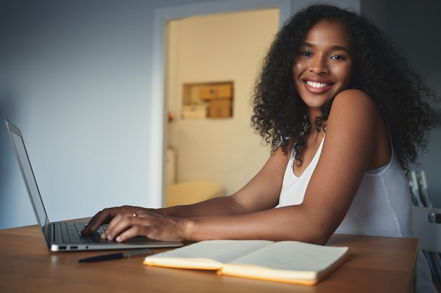 Concepto de personas, estilo de vida, tecnología moderna, trabajo y comunicación. Atractiva joven autónoma de piel oscura con cabello rizado con mirada complacida, disfrutando del trabajo distante, usando laptop genérica