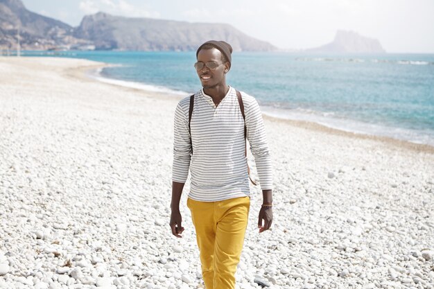 Concepto de personas, estilo de vida, turismo, viajes y vacaciones. Atractivo alegre joven mochilero afroamericano disfrutando de las vacaciones de verano en la playa junto al mar