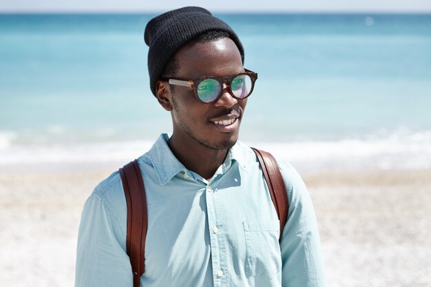 Concepto de personas, estilo de vida, viajes, aventura, vacaciones y turismo. Turista europeo negro de moda en ropa de moda que se relaja en la playa en un día soleado de verano, caminando solo por la orilla del mar